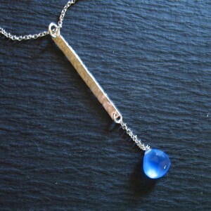 May include: A silver chain necklace with a hammered silver bar pendant and a blue gemstone drop.