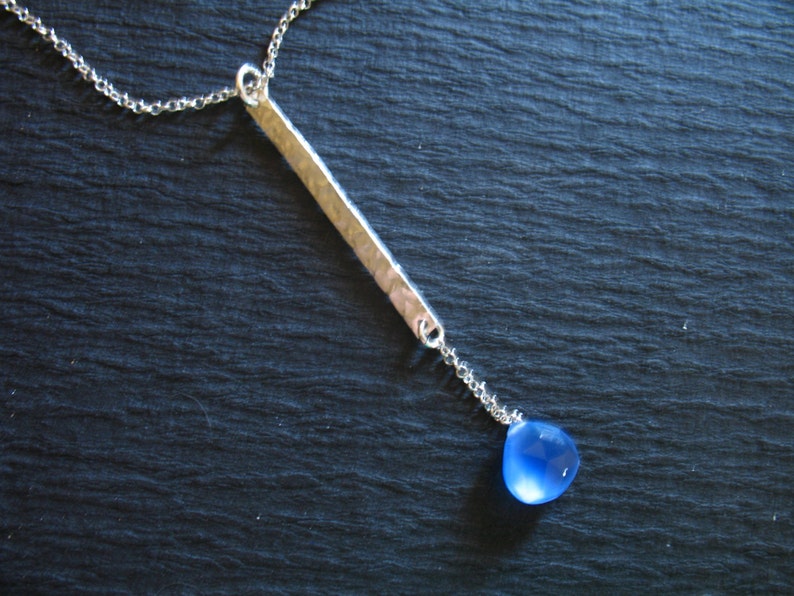 May include: A silver chain necklace with a hammered silver bar pendant and a blue gemstone drop.