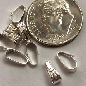 May include: A close-up of a group of silver-toned pendant bails. The bails are oval-shaped and have a smooth, polished finish. The bails are arranged in a random pattern on a white surface. A US dime is included for size comparison.