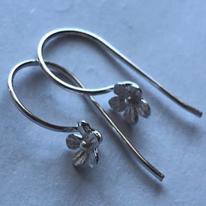 Pode incluir: Um par de brincos de flor pendentes de prata. Os brincos são feitos de um fio fino com um pequeno pingente de flor na ponta.