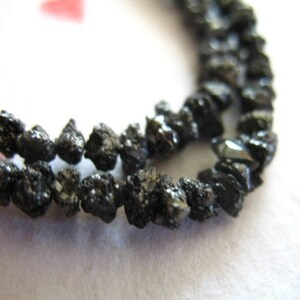 Puede incluir: Una hebra de cuentas de diamante negro, cada cuenta tiene una forma irregular y tosca. Las cuentas est&aacute;n enhebradas para formar un collar o una pulsera.