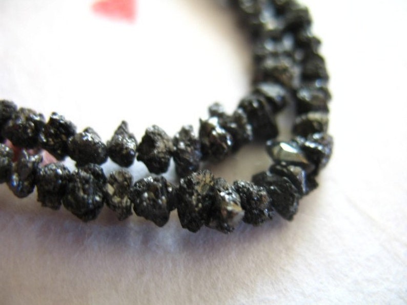 Puede incluir: Una hebra de cuentas de diamante negro, cada cuenta tiene una forma irregular y tosca. Las cuentas est&aacute;n enhebradas para formar un collar o una pulsera.