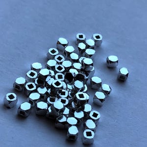 May include: A pile of silver-colored metal beads with a square hole in the center of each bead. The beads are hexagonal in shape.