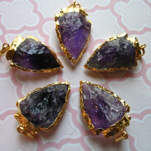 May include: Five amethyst arrowhead pendants with gold-plated edges. The arrowheads are all different sizes and shapes, but they are all a deep purple color.