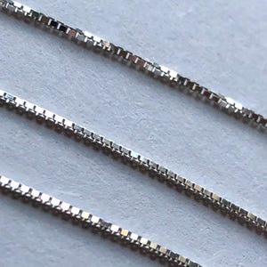 May include: Three silver box chains, each with a different length, are laid out on a white surface. The chains are made of small, square links.