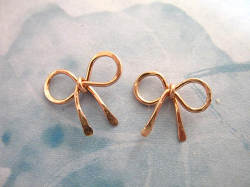 May include: A pair of rose gold bow earrings. The bows are made of wire and have a hammered texture.