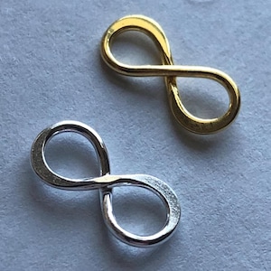 Könnte beinhalten: Zwei Unendlichkeits-Anhänger, einer in Silber und einer in Gold. Die Anhänger sind aus Metall und etwa 2,5 cm groß.