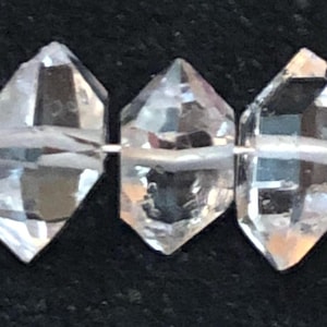 May include: A strand of six clear quartz crystal beads, each with a faceted, diamond-shaped design. The beads are strung on a thin, white thread.