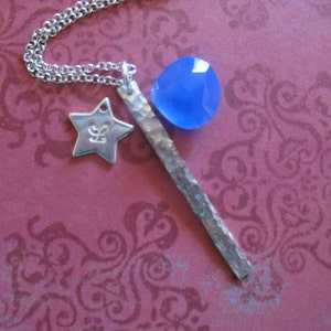 May include: A silver necklace with a blue gemstone pendant, a star charm, and a hammered silver bar. The necklace is on a pink background with a floral pattern.