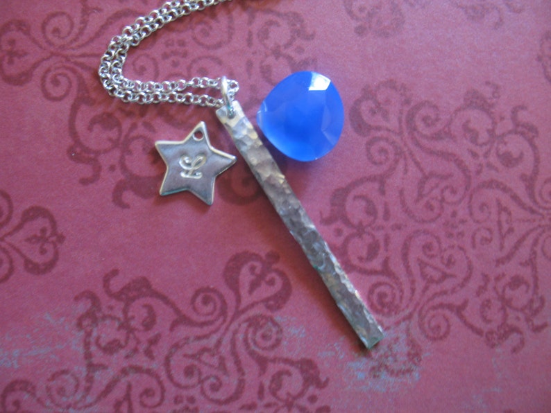 May include: A silver necklace with a blue gemstone pendant, a star charm, and a hammered silver bar. The necklace is on a pink background with a floral pattern.