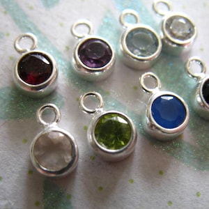 May include: Assortment of nine silver charm pendants with different colored gemstones. The gemstones include clear, green, blue, black, red, and purple.