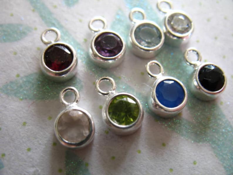 May include: Assortment of nine silver charm pendants with different colored gemstones. The gemstones include clear, green, blue, black, red, and purple.