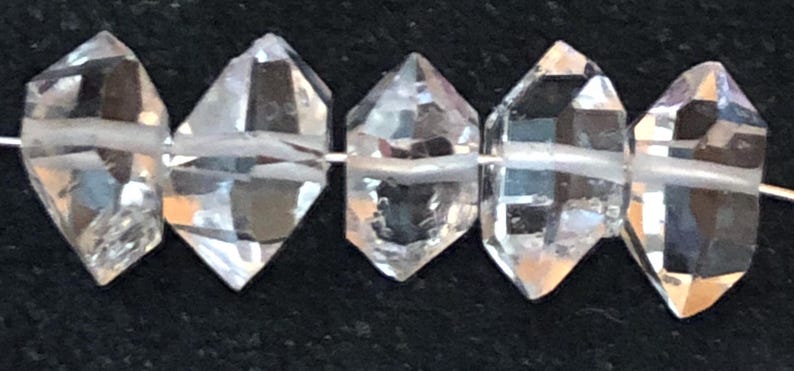 May include: A strand of six clear quartz crystal beads, each with a faceted, diamond-shaped design. The beads are strung on a white thread.
