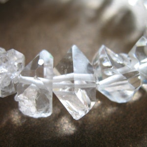 May include: A close-up of a crystal bracelet made of clear, faceted quartz stones strung on white thread. The stones are various sizes and shapes, with some having a more rounded appearance.