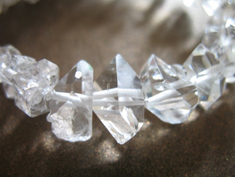 May include: A close-up of a crystal bracelet made of clear, faceted quartz stones strung on white thread. The stones are various sizes and shapes, with some having a more rounded appearance.