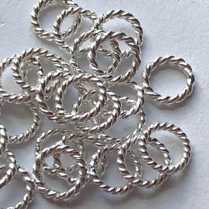 May include: A pile of silver jump rings with a twisted rope design. The rings are small and delicate, and they are perfect for jewellery making.
