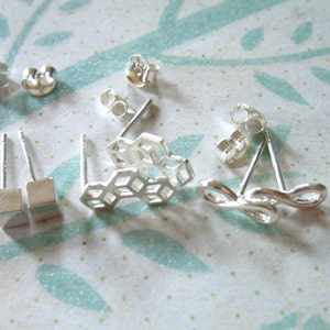 May include: A collection of silver stud earrings in various designs. Includes pairs with square studs, honeycomb shapes, and infinity loop designs. Earring backs are also visible. The earrings are displayed on a light blue and white patterned surface.