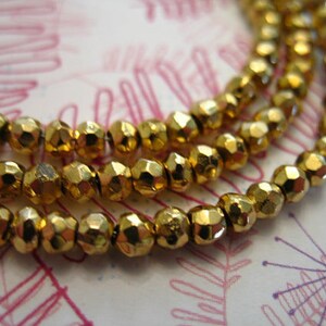 May include: A close-up of a gold-colored beaded necklace. The beads are faceted and have a metallic sheen. The necklace is layered and appears to be made of multiple strands of beads.