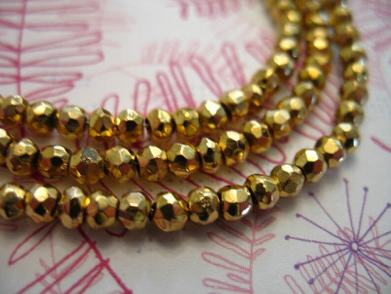 May include: A close-up of a gold-colored beaded necklace. The beads are faceted and have a metallic sheen. The necklace is layered and appears to be made of multiple strands of beads.
