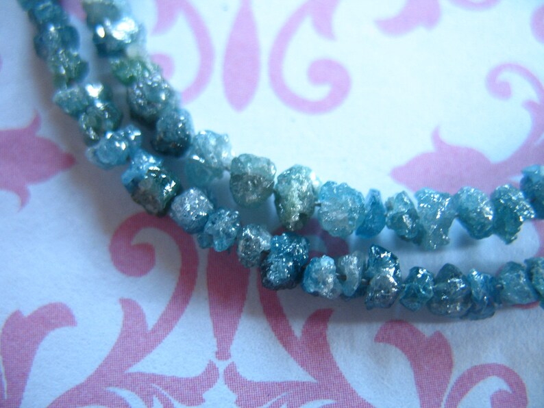 Puede incluir: Una hebra de diamantes en bruto azules y verdes, tambi&eacute;n conocidos como diamantes en bruto, sobre un fondo blanco con un patr&oacute;n floral rosa.