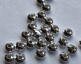 Può includere: Un gruppo di perline rotonde color argento con un foro al centro. Le perline sono sparse su una superficie bianca.