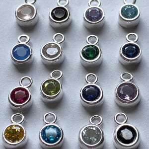 May include: Assortment of 16 silver charm pendants with different colored gemstones. Each pendant features a round gemstone in a silver setting with a loop for attaching to a chain or bracelet.