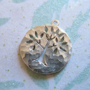 May include: Silver charm in the shape of a tree with a hammered texture. The tree has a thick trunk and many branches with leaves.