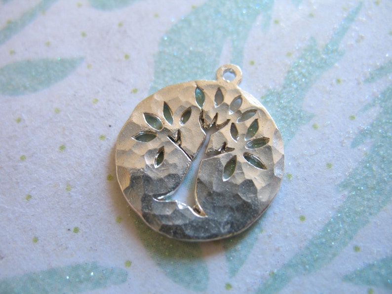 May include: Silver charm in the shape of a tree with a hammered texture. The tree has a thick trunk and many branches with leaves.