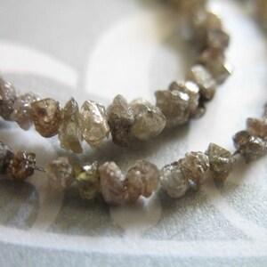 Puede incluir: Una hebra de diamantes en bruto marrones y grises enhebrados en un alambre. Los diamantes tienen una forma irregular y un aspecto natural y sin pulir.