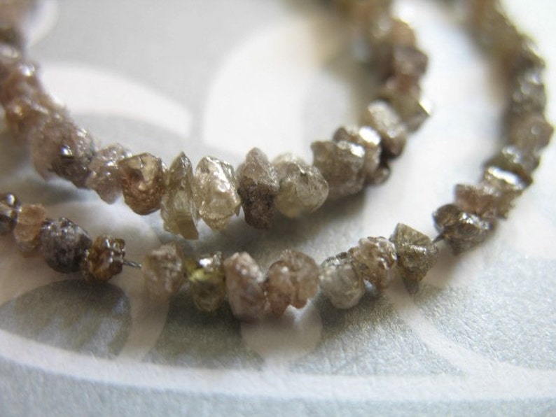 Puede incluir: Una hebra de diamantes en bruto marrones y grises enhebrados en un alambre. Los diamantes tienen una forma irregular y un aspecto natural y sin pulir.
