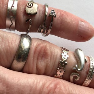 May include: A hand wearing a variety of silver rings, including a wave ring, a shell ring, an arrow ring, a band ring, a flower ring, a dolphin ring, and a textured ring.