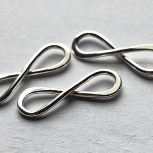 Könnte beinhalten: Drei silberne Charms in Form eines Unendlichkeitszeichens. Die Charms sind glatt und glänzend und perfekt, um jedem Schmuckprojekt einen Hauch von Eleganz zu verleihen.