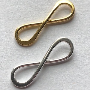 Pode incluir: Dois pingentes com o símbolo do infinito, um dourado e outro prateado. Os pingentes são feitos de metal e medem aproximadamente 2,5 cm de comprimento.