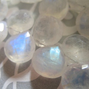 May include: A collection of iridescent white moonstone beads with a pearlescent sheen. The beads are faceted and have a smooth, polished finish.