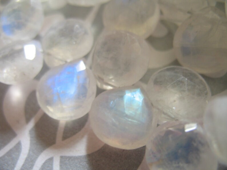 May include: A collection of iridescent white moonstone beads with a pearlescent sheen. The beads are faceted and have a smooth, polished finish.