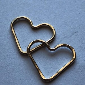 May include: Two gold heart-shaped hoops, one slightly larger than the other, are stacked on top of each other. The hoops are made of a hammered metal, giving them a textured look.