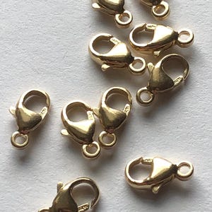 May include: A collection of gold-colored lobster claw clasps, each with a spring-loaded mechanism and a small loop for attaching to jewelry. The clasps are arranged on a light-colored surface, showcasing their shiny finish.