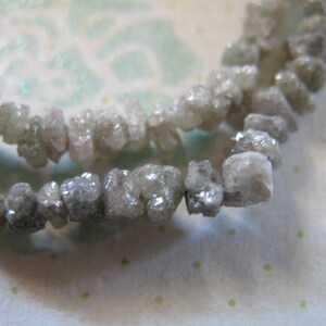 Puede incluir: Una hebra de diamantes grises en bruto enhebrados como un collar. Los diamantes son de forma irregular y tienen un aspecto natural sin pulir.