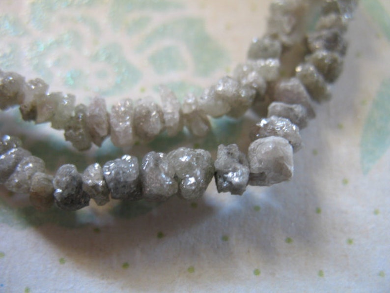Puede incluir: Una hebra de diamantes grises en bruto enhebrados como un collar. Los diamantes son de forma irregular y tienen un aspecto natural sin pulir.