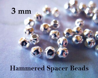 Op de afbeelding: Een close-up van zilveren gehamerde spacer kralen, 3 mm in grootte. De kralen zijn verspreid over een lichtblauw oppervlak.