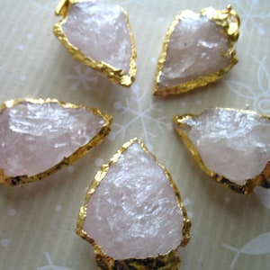 May include: Five rose quartz arrowhead pendants with gold-plated edges. The arrowheads are all different shapes and sizes.