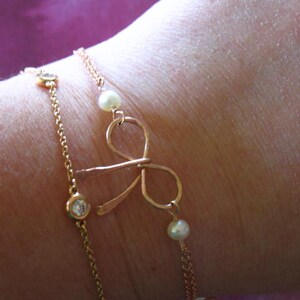 May include: A gold chain bracelet with a bow charm and small pearl accents. The bracelet is layered with a second chain with small clear stones.