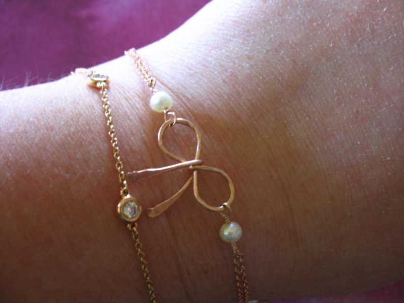 May include: A gold chain bracelet with a bow charm and small pearl accents. The bracelet is layered with a second chain with small clear stones.