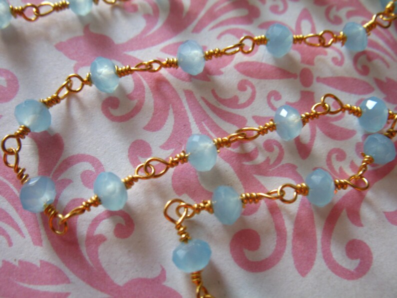 May include: A delicate gold chain necklace with light blue gemstone beads. The necklace is made with small, round beads and is strung on a thin gold chain.