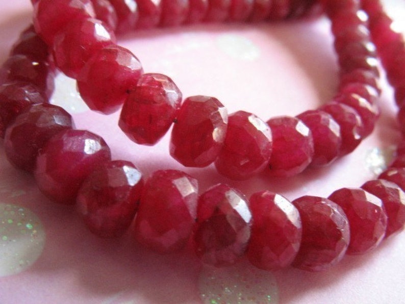 Puede incluir: Una hebra de cuentas de rub&iacute; rojo intenso, facetadas y pulidas, ensartadas para formar un collar o pulsera. Las cuentas est&aacute;n dispuestas en primer plano, mostrando su rico color y sus intrincadas facetas.