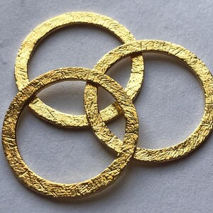 Op de afbeelding: Drie gouden metalen ringen die in elkaar gehaakt zijn. De ringen hebben een getextureerd oppervlak.