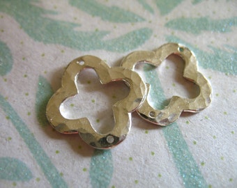 May include: Two silver-colored, hammered-metal clover-shaped charms. Each charm has four rounded lobes and a small hole for stringing. The charms are displayed on a textured, light-colored surface with a subtle green and white pattern.