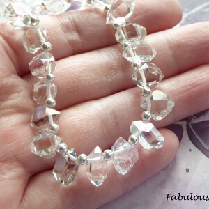 May include: A strand of clear quartz crystal beads with silver accents. The beads are faceted and have a diamond-like shape. The beads are strung on a white cord.