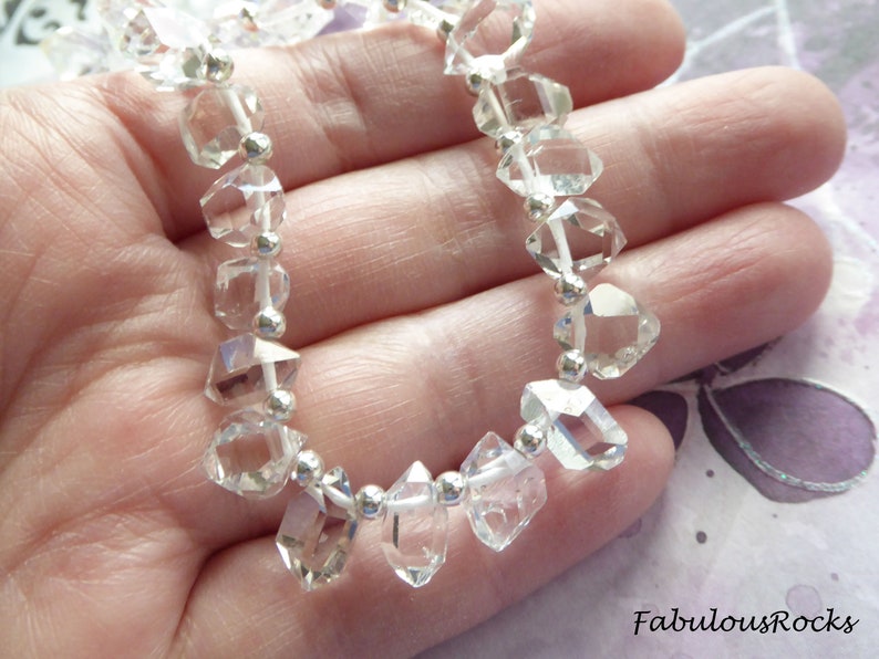 May include: A strand of clear quartz crystal beads with silver accents. The beads are faceted and have a diamond-like shape. The beads are strung on a white cord.