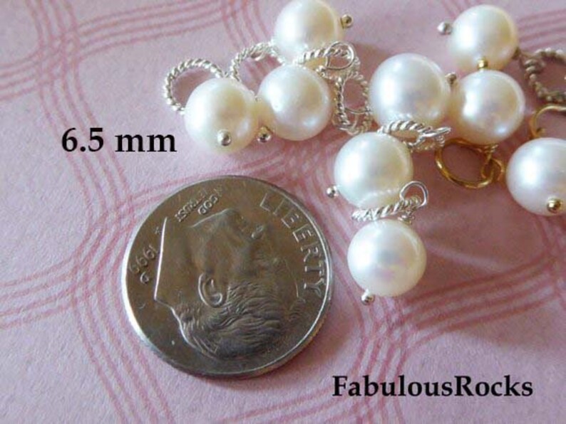 May include: A close-up of a group of white pearl beads with silver and gold accents. The pearls are 6.5 mm in diameter and are arranged in a cluster. The beads are on a pink surface with a silver coin in the background.